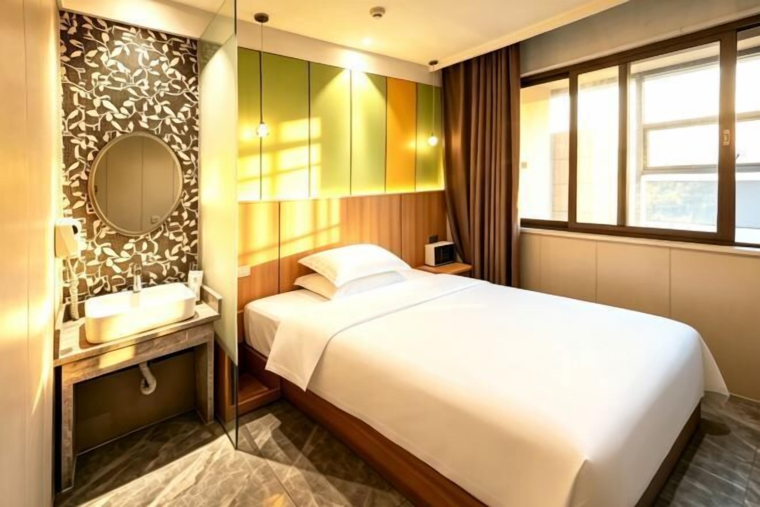 A hotel room with a bed, a bedside table, a mirror, a sink, and a window with curtains.
