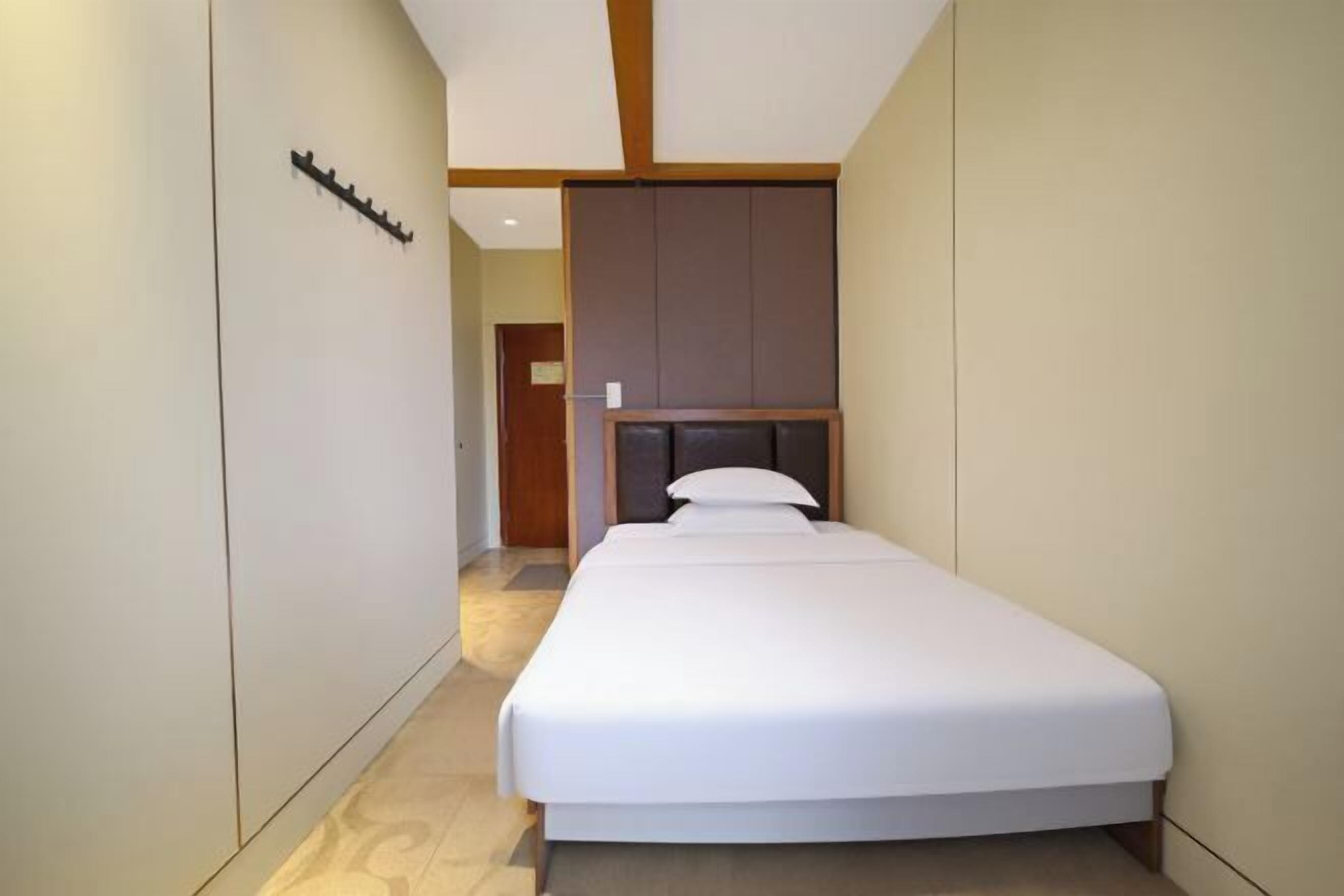A narrow hotel room with a bed, a headboard, and a coat rack.