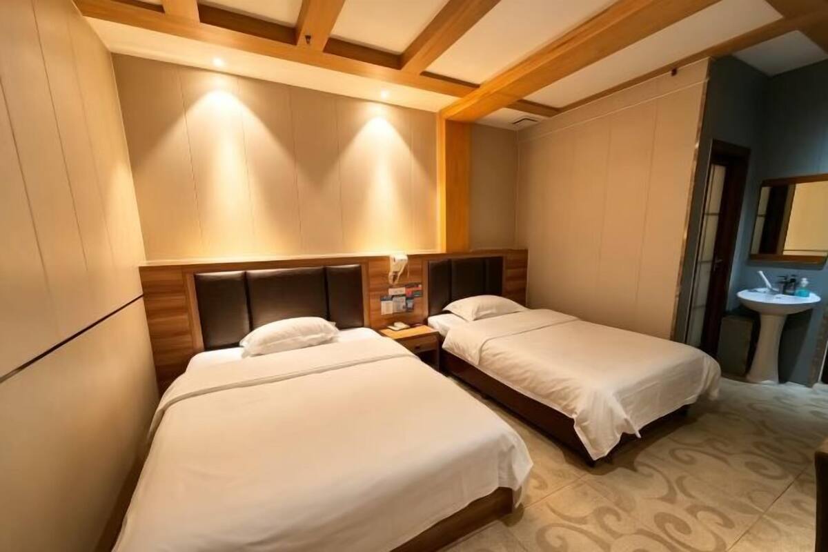 Habitación de hotel con dos camas, cabeceros de madera, una mesita de noche con lámpara, una pequeña mesa redonda con espejo y un estante de pared con libros.