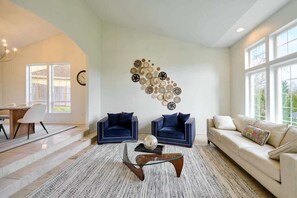 A living room with a sofa, two armchairs, a coffee table, a wall art piece, and a dining area with a table and chairs.
