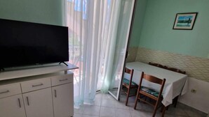 A small, clean room with a TV on a cabinet, a dining table with chairs, and a sliding glass door to a balcony.