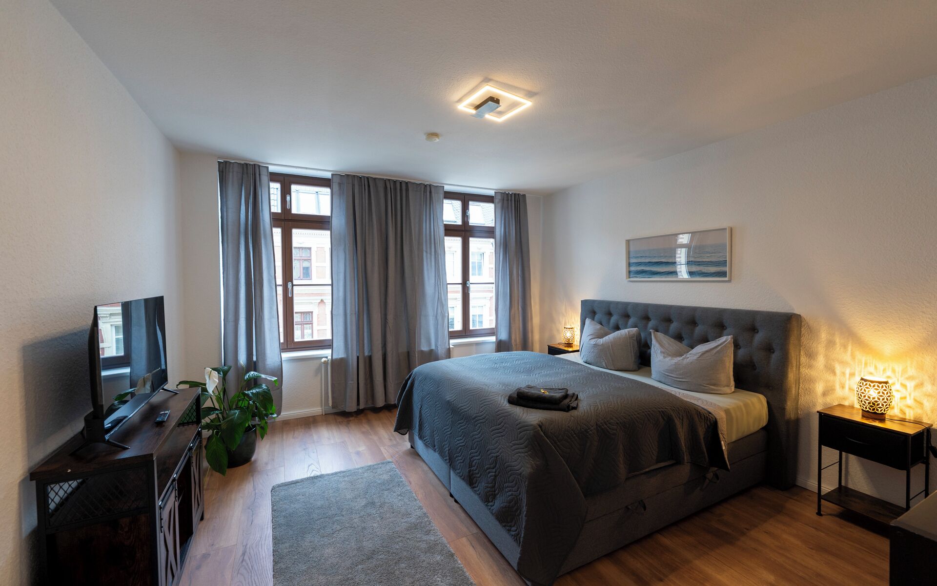 A modern bedroom with a large bed, a desk, a TV, and a window with curtains.