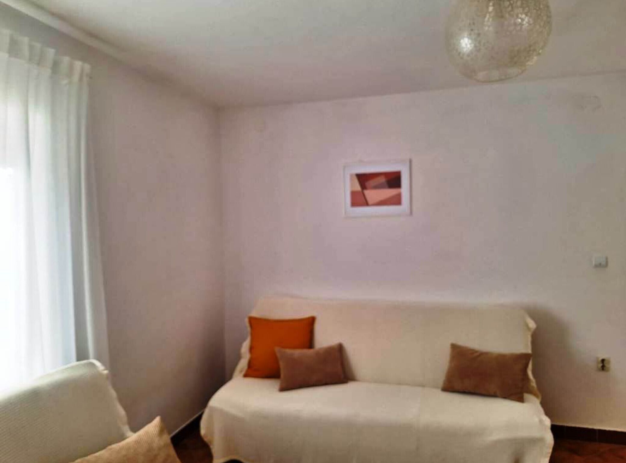A room with a white sofa, orange and brown cushions, a framed picture on the wall, and a hanging light fixture.