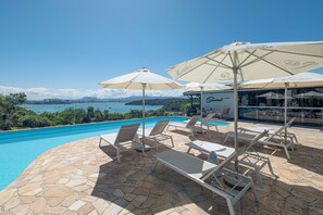 Zona de piscina amb gandules i ombrelles, amb vista a un mar blau clar i muntanyes llunyanes.