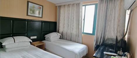 A hotel room with two single beds, a desk with a chair, a window with curtains, a picture on the wall, and an air conditioning unit.
