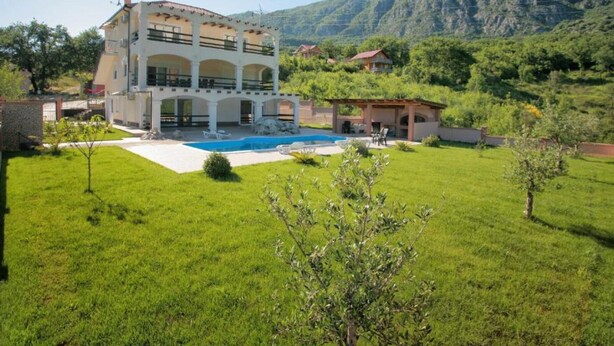 A two-story house with a swimming pool, surrounded by a lush green lawn and mountains in the background.