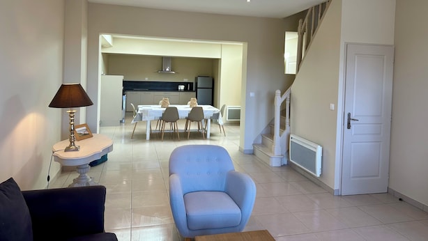 A modern living room with a sofa, a dining table with chairs, a kitchen in the background, and a staircase.