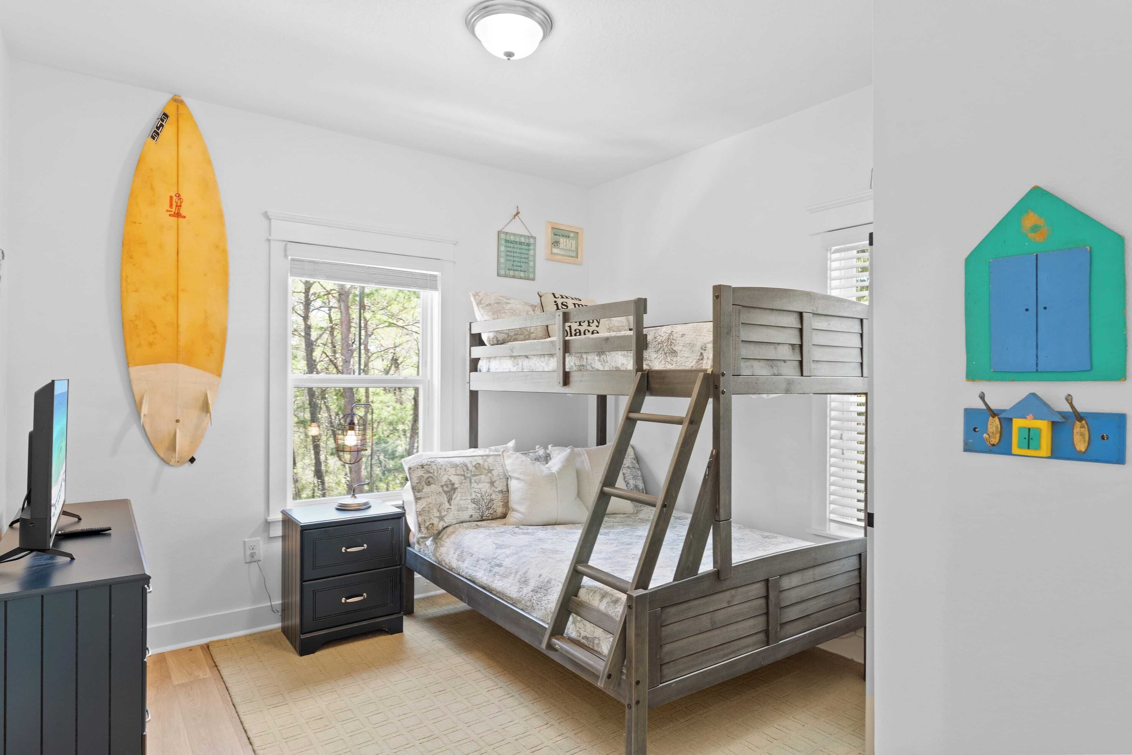 Un dormitorio con literas, una tabla de surf, un escritorio y una ventana con vista a árboles.