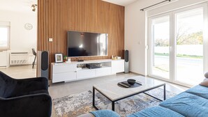 A modern living room with a large flat-screen TV, wooden panel wall, white entertainment unit, and a glass door leading outside. - Holiday Home Iva - Three Bedroom Holiday Home (Šibenik)