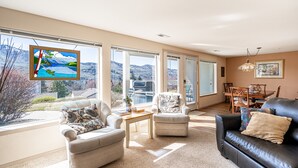 A spacious living room with a large window offering a mountain view, a black leather sofa, a beige armchair, and a wooden coffee table. - Mountain View Retreat - Condo in Chelan (Chelan)