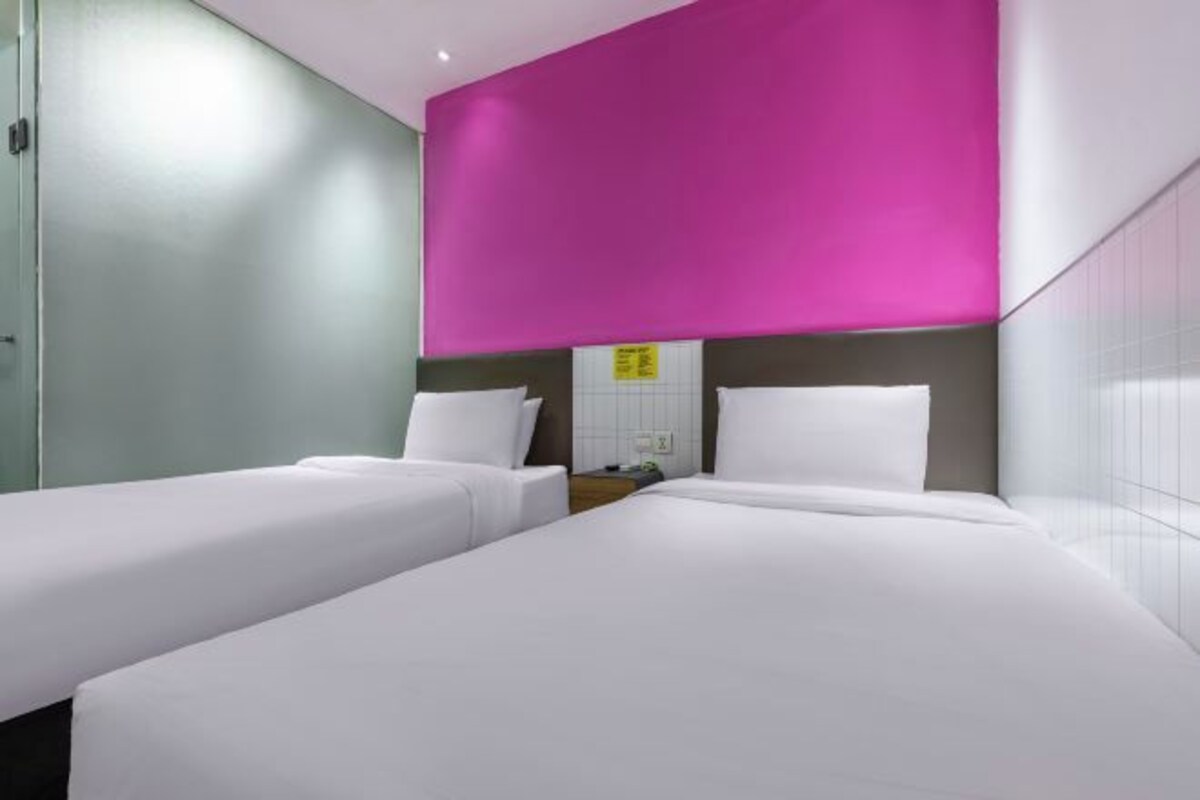 A hotel room with two single beds, a glass partition, and a bright pink accent wall.