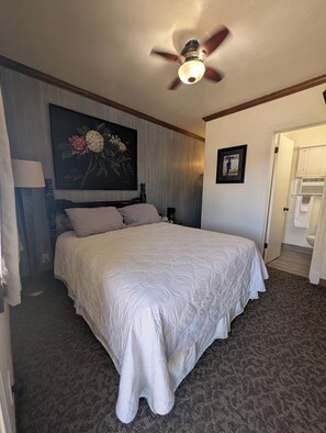 A hotel room with a large bed, a ceiling fan, a painting, a framed picture, and a bathroom door. - Squaw Valley Motel Room #4 (Yokuts Valley)