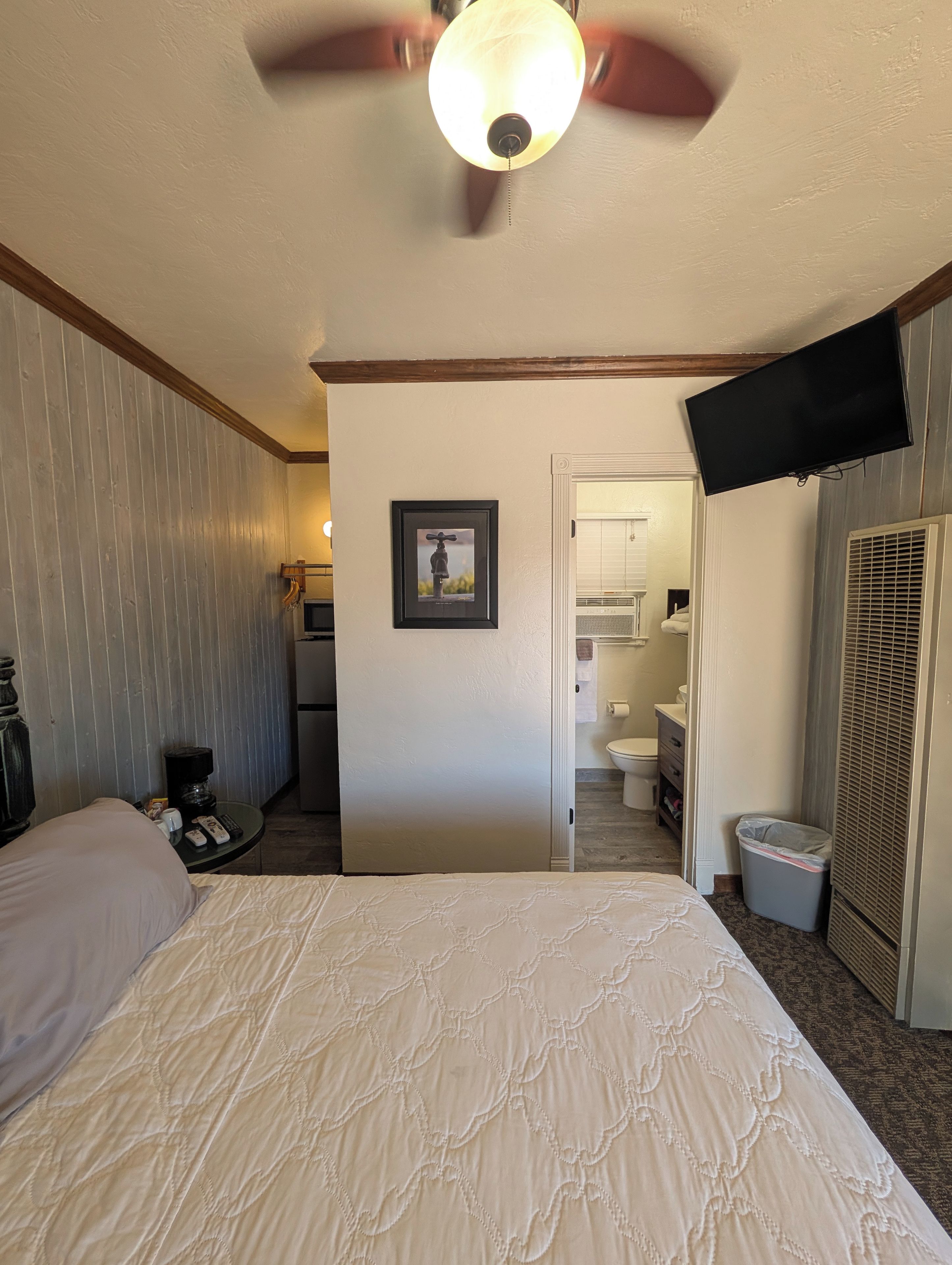 A hotel room with a bed, a ceiling fan, a television, a picture frame, and a bathroom visible through an open door.