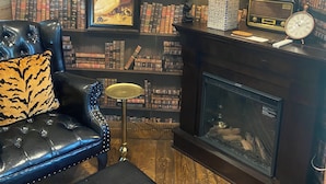 A cozy reading nook with a leather armchair, a small side table, a bookshelf filled with books, a framed painting, and a fireplace.