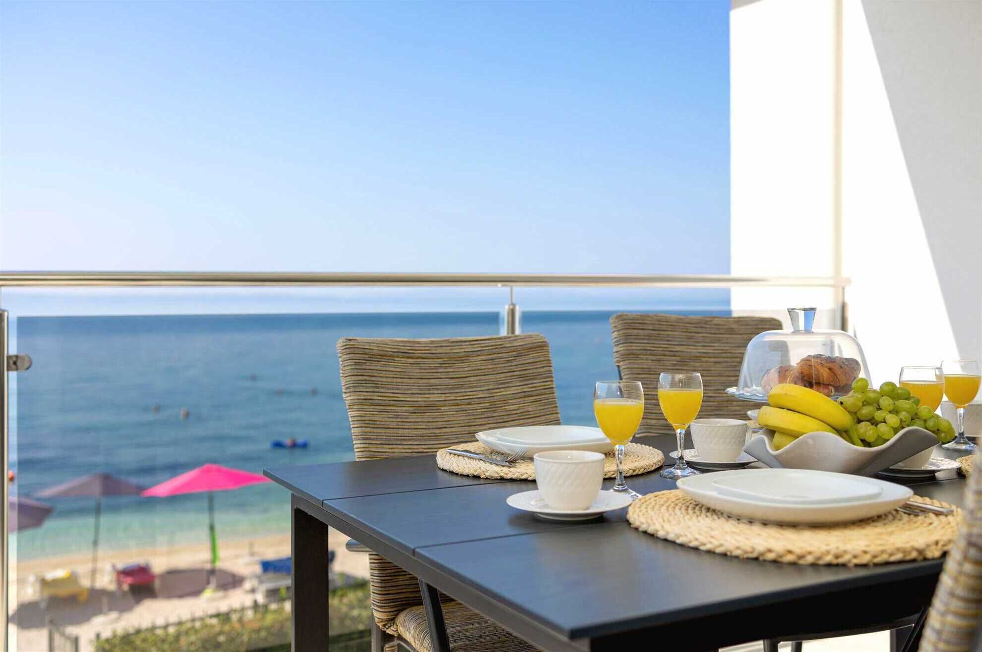 A balcony table with breakfast items, a view of the sea, and beach umbrellas in the background.