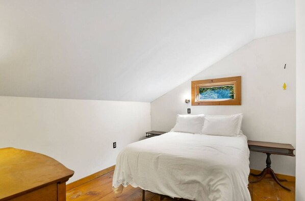 A bedroom with a sloped ceiling, a bed with white bedding, a wooden nightstand, and a painting on the wall.