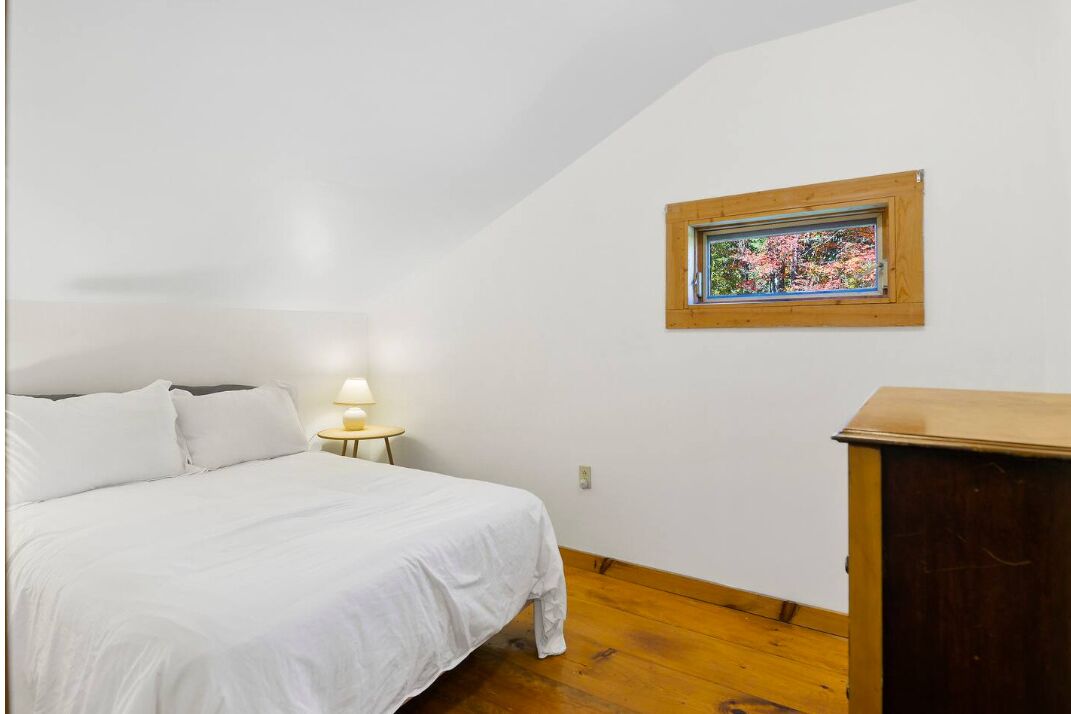 A bedroom with a bed, a nightstand with a lamp, a wooden dresser, and a window with a view of trees.