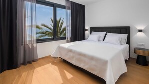 A hotel room with a large bed, a nightstand, a wall-mounted lamp, and a view of palm trees through the window.