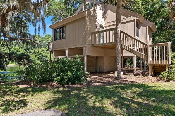 A two-story house with a deck and a covered porch, surrounded by trees and a pond. - 67 Night Heron | Lagoon Views + Loft + Pool (Hilton Head Island)