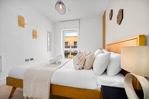Une chambre à coucher moderne avec un cadre de lit en bois, du linge de lit blanc et une vue sur la ville par une grande fenêtre.