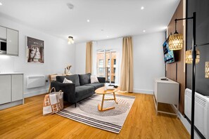 A modern living room with a sofa, a coffee table, and a TV. There is a window with curtains, a radiator, and a wooden floor. - Stylish 2 Bed Apartment - 2 Bathrooms - Balcony (West Midlands)