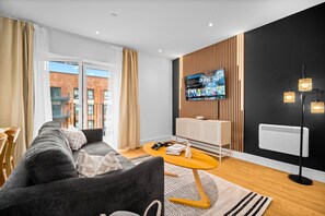 A modern living room with a grey sofa, a wooden sideboard, a TV mounted on a wooden panel wall, and a floor lamp. - Stylish 2 Bed Apartment - 2 Bathrooms - Balcony (West Midlands)