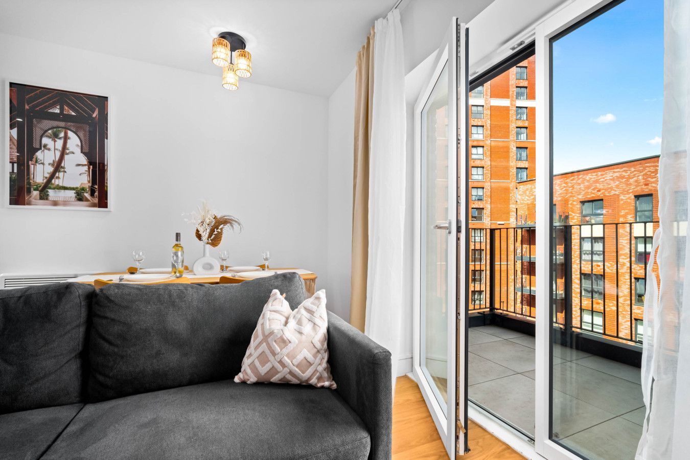 Un salon moderne avec un canapé gris, une table à manger dressée pour quatre personnes, une grande fenêtre donnant sur la ville et un luminaire au plafond.