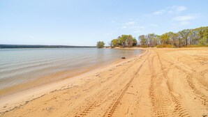 A sandy beach with tire tracks leading to the water, a clear sky, and trees in the background. - Walk to Beach: Eufaula 'Fisherman’s Delight' Home (Eufaula)