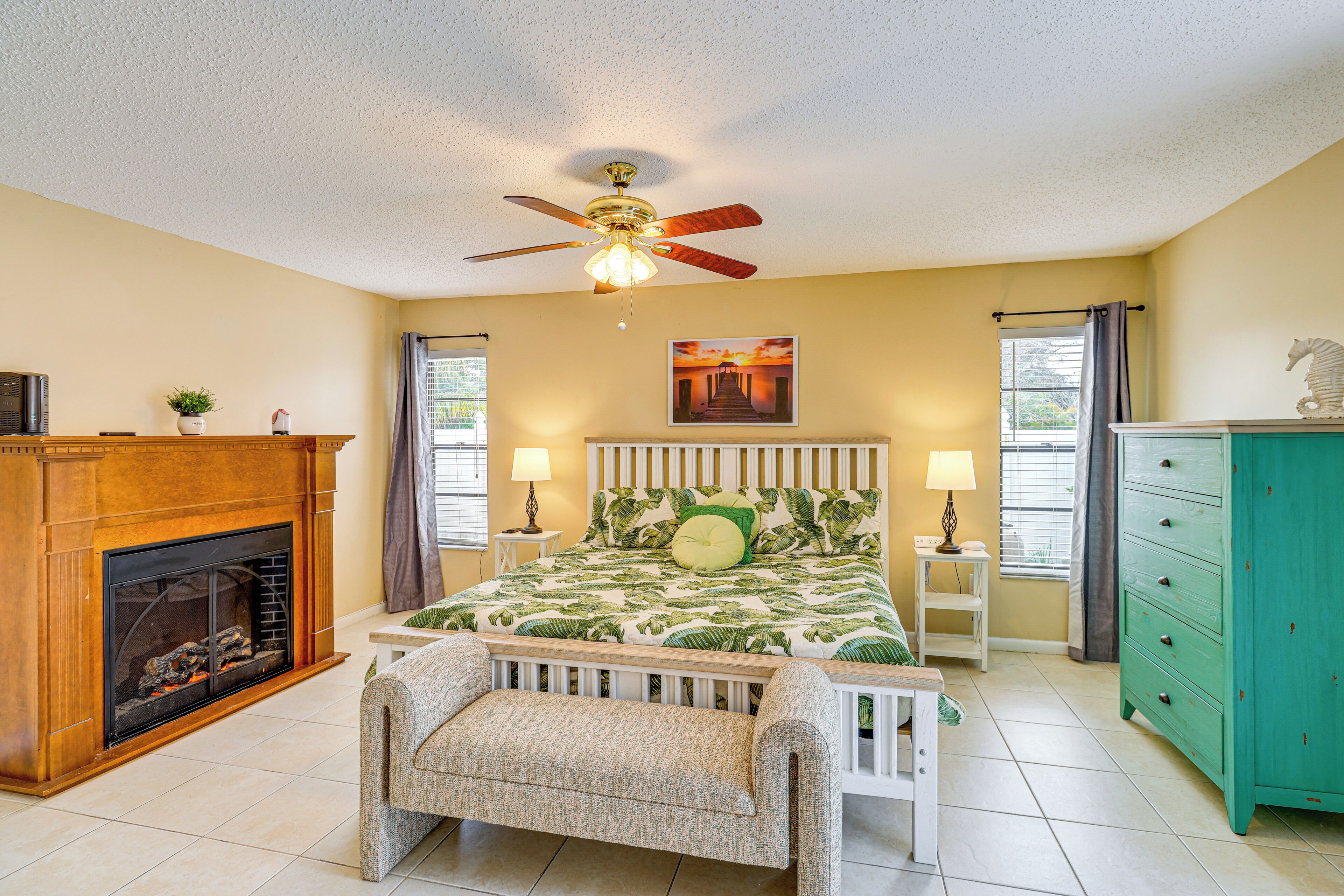 A bedroom with a fireplace, a bed with a green leaf pattern, a ceiling fan, and a green dresser.