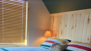 Ein Schlafzimmer mit Bett, Fenster mit Jalousien, einer Lampe und einem hölzernen Kopfteil.