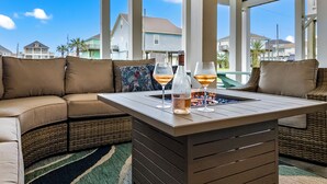 A modern outdoor seating area with a cushioned sofa, a wooden table with two glasses of wine, and a bottle of wine. - Family Friendly-Sleeps 10-BBQ-EV-Game Rm-Walk to Beach-Fire Table-Fully Stocked (Crystal Beach)