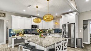 A modern kitchen with white cabinets, a large island with bar stools, stainless steel appliances, and decorative lighting. - Family Friendly-Sleeps 10-BBQ-EV-Game Rm-Walk to Beach-Fire Table-Fully Stocked (Crystal Beach)