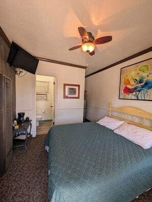 A hotel room with a bed, a ceiling fan, a painting, a desk with a coffee maker, and a door leading to a bathroom. - Squaw Valley Motel Room #5 (Yokuts Valley)