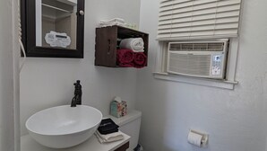 Een badkamer met een wit wastafel, een donker houten wastafelmeubel, een spiegel met verlichting, een toilet en een raam met jaloezieën.