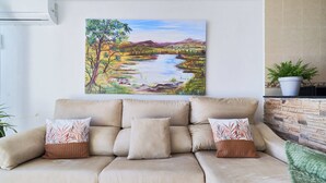 A beige sofa with two decorative pillows, a painting of a landscape, and a potted plant on a tiled wall.