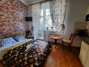 A room with a brick wall, a bed with patterned bedding, a small round table, a chair, a window with patterned curtains, and a television on the wall.