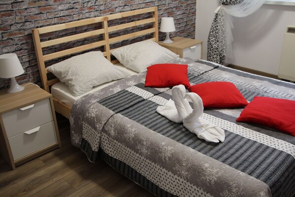 Een net opgemaakt bed met een gestreept dekbedovertrek, twee kussens en een handdoek. Een houten hoofdbord, een nachtkastje met een lamp en een klein ladekastje. De kamer heeft een bakstenen muur en een raam met een dunne gordijn.