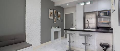 Una cocina moderna con encimera de mármol blanco, microondas integrado y refrigerador. Hay tres taburetes de bar y una pequeña mesa con un vaso encima.