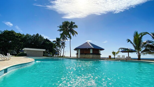 A clear blue pool with a small pavilion, palm trees, and lounge chairs. - Enjoy the Wonderful Ocean & Pool View from This 2BDR Apartment in Juan Dolio (Guayacanes)