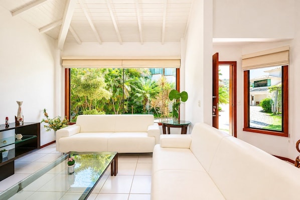A spacious living room with a white sofa, a glass coffee table, and a view of lush greenery outside.
