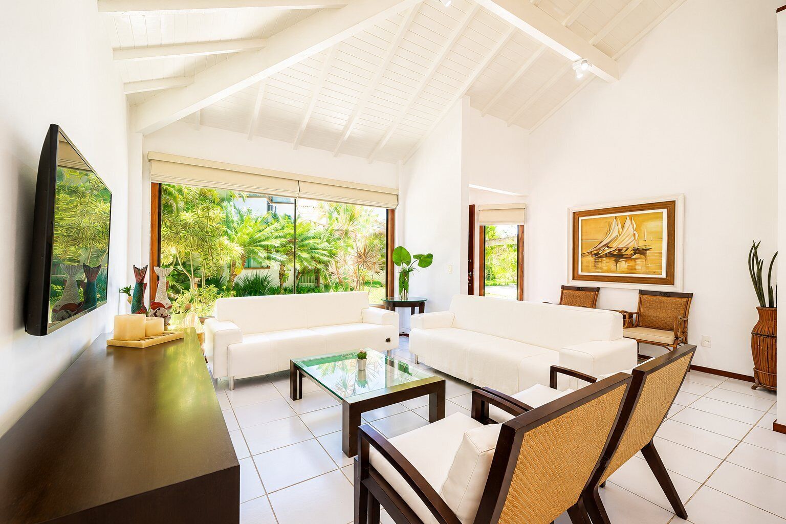 A spacious living room with a white sofa, a glass coffee table, and a large window offering a view of greenery.