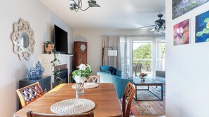 Un coin repas avec une table en bois, des chaises et un vase de fleurs. Il y a une horloge au mur et un téléviseur posé sur un meuble.
