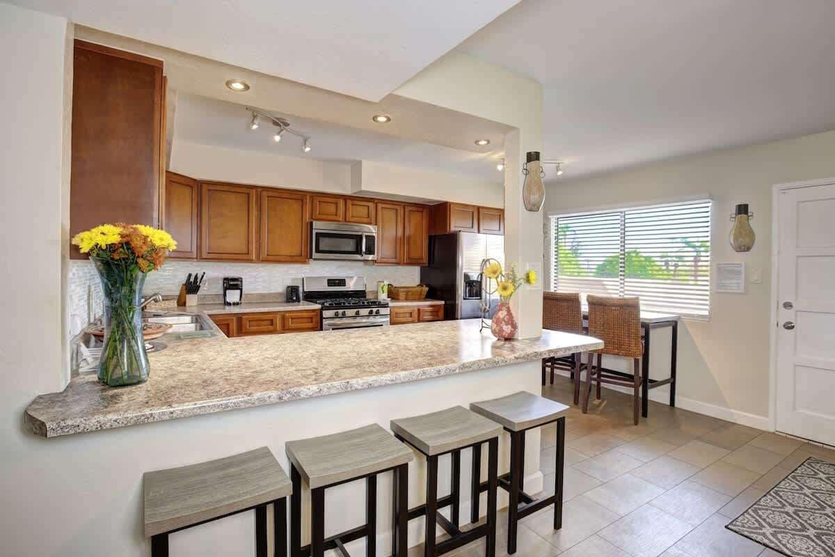 A modern kitchen with wooden cabinets, a granite countertop, a central island with bar stools, and a dining area with wicker chairs.