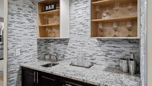 A kitchen with a granite countertop, a sink, a backsplash with a stone-like pattern, and wooden shelves with wine glasses. - Modern, remodeled, pool table in Wellington! (Wellington)