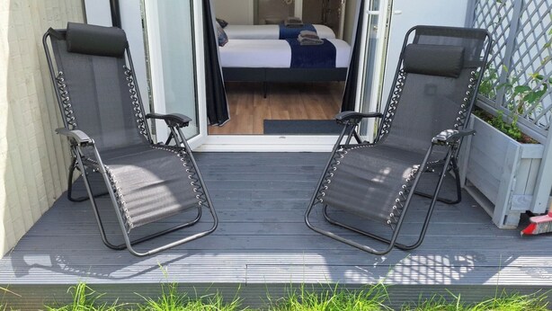 Dos sillas plegables negras sobre una terraza de madera, con una cama visible a través de una puerta abierta.
