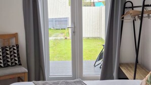 Habitación de hotel con cama, cabecero de madera, manta gris, perchero de madera y vista al exterior.