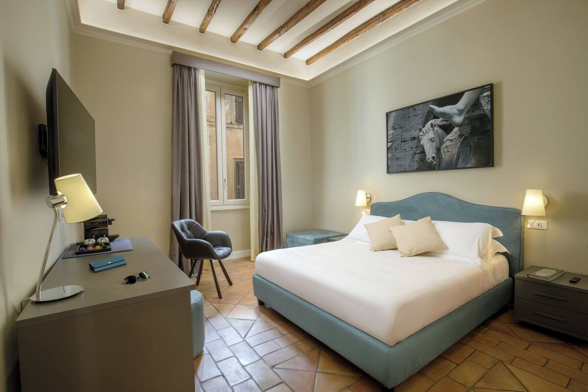 A hotel room with a bed, desk, chair, and a painting on the wall.