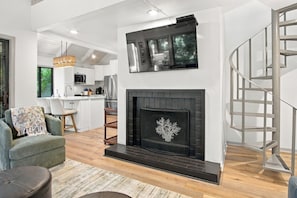 A modern living room with a fireplace, a staircase, and a kitchen in the background.