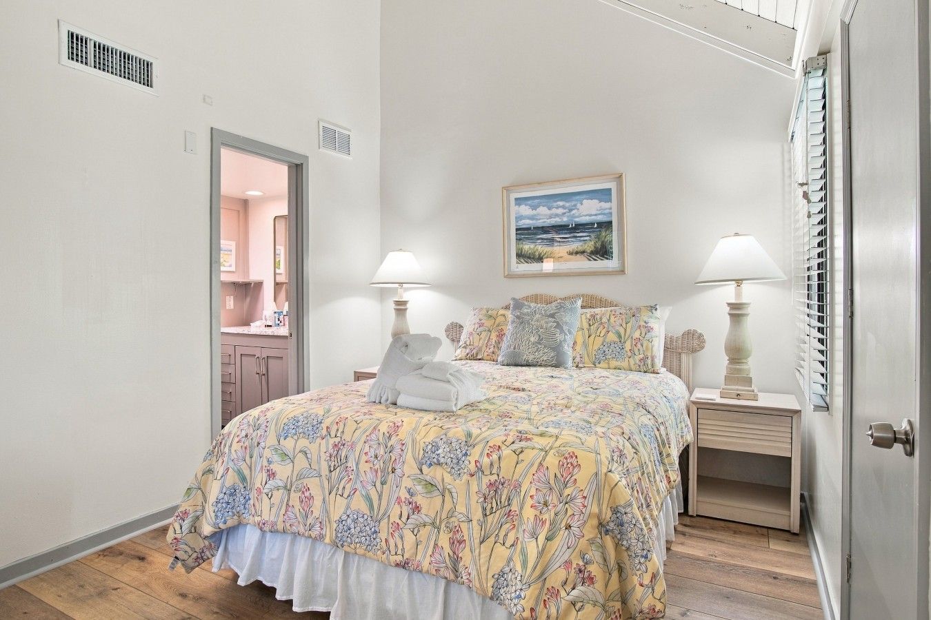 A bedroom with a bed, two bedside tables, a nightstand, a door, a window with blinds, and a painting on the wall.