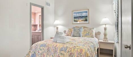 A bedroom with a bed, two bedside tables, a nightstand, a door, a window with blinds, and a painting on the wall.